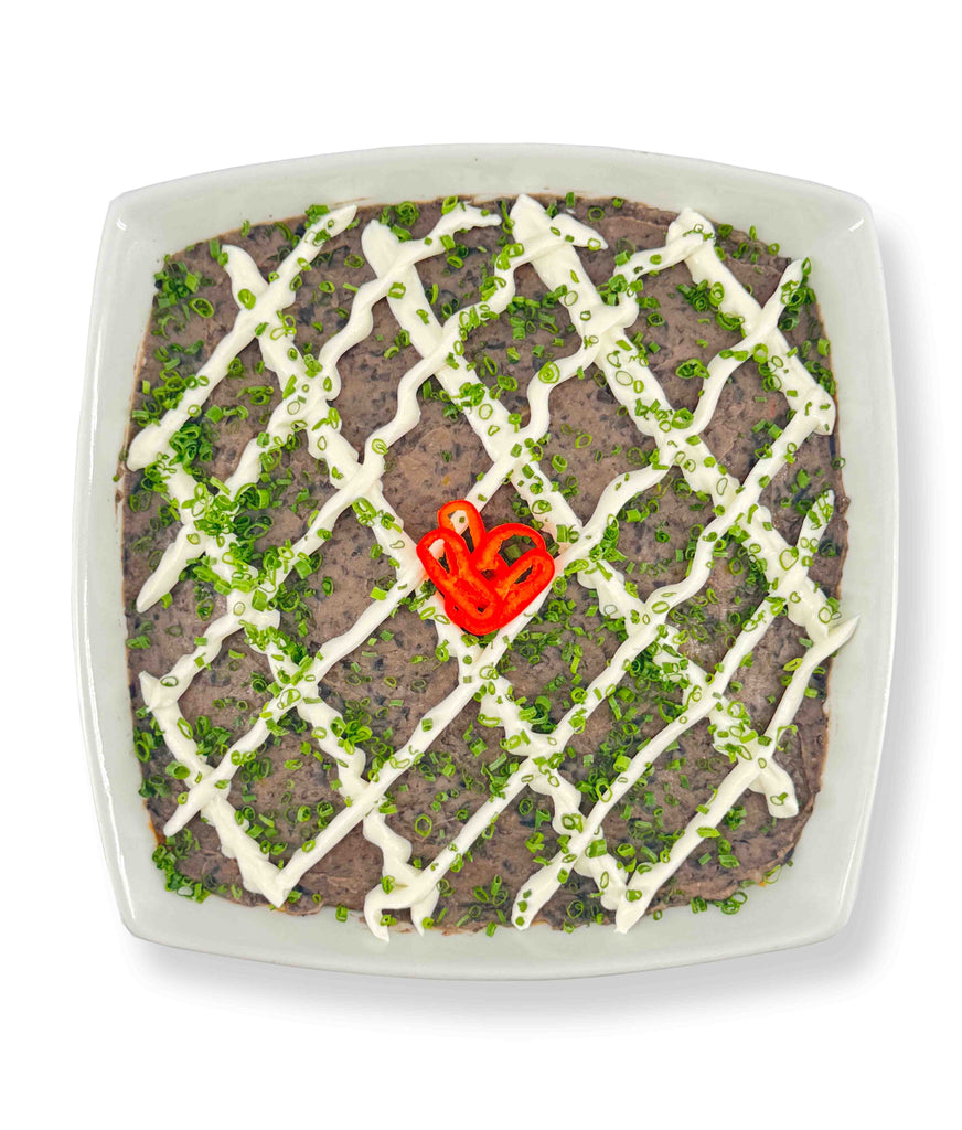 A square dish of 6 Layer Dip topped with Whitestone Cheddar, a mayonnaise drizzle, green onions, and a red chili pepper slice in the center. Serve with guacamole and corn tortillas for a complete flavor experience.