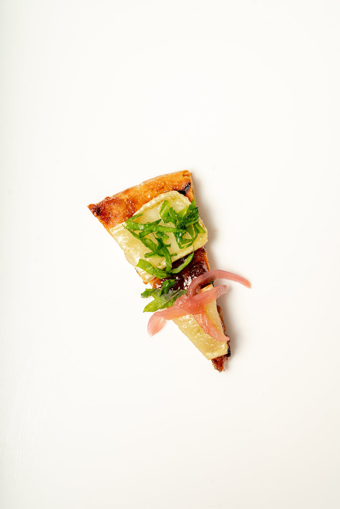 A Fig And Brie Naan Flatbread topped with melted cheese, baby arugula, and pickled red onions on a plain white background.