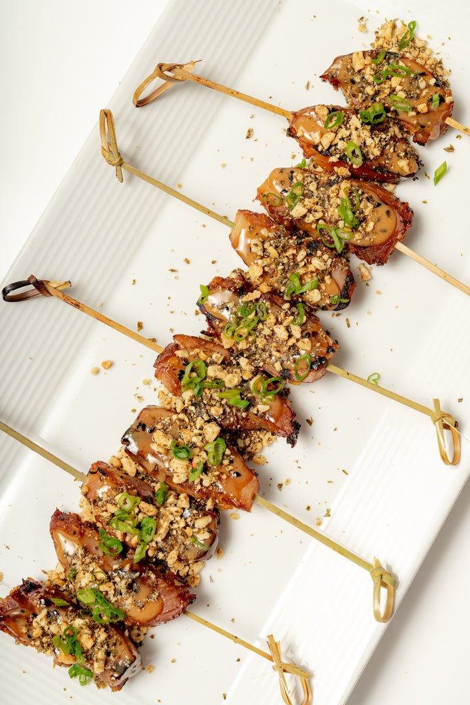 Grilled Chicken Satay—four skewers topped with chopped nuts and green onions—are served in a row on a rectangular white plate with our signature Peanut Sesame Dipping Sauce.
