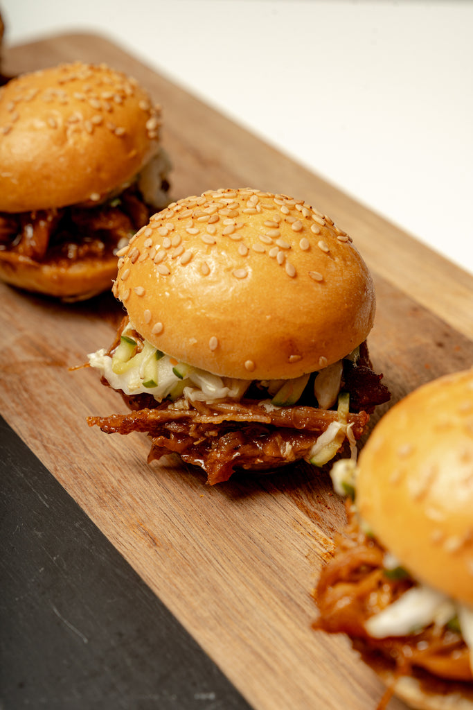 Three Hot Honey Pulled Chicken Sliders with pickle slaw are served on mini brioche buns, arranged on a wooden serving board.
