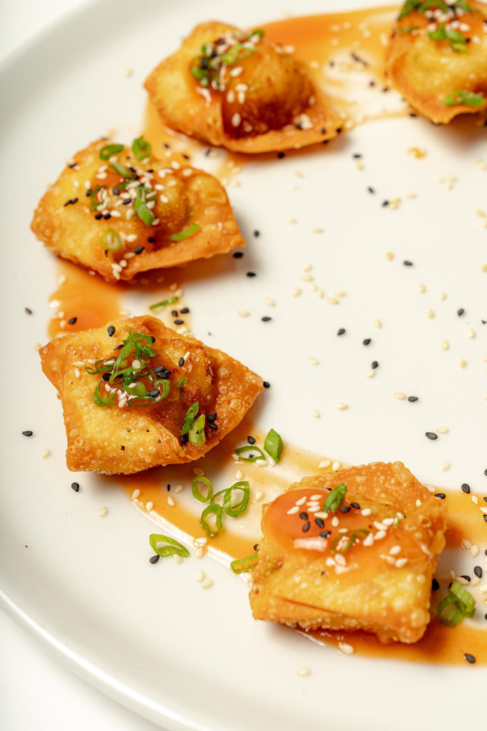 Five crispy Lobster Rangoons filled with lobster and cream cheese are topped with green onions, sesame seeds, and sauce, and served on a white plate.