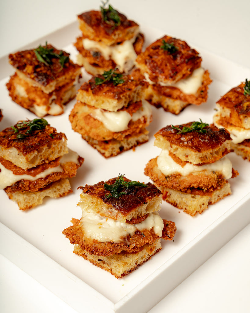 Tray of Mini Chicken Parm On Foccacia—stacked mini sandwiches with golden brown bread, melted cheese, and basil pesto—arranged neatly on a white square plate.
