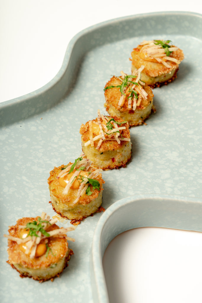 Five small, round Mini Green Curry Crabcakes topped with shredded cheese and herbs are arranged in a curved line on a light blue, textured platter.