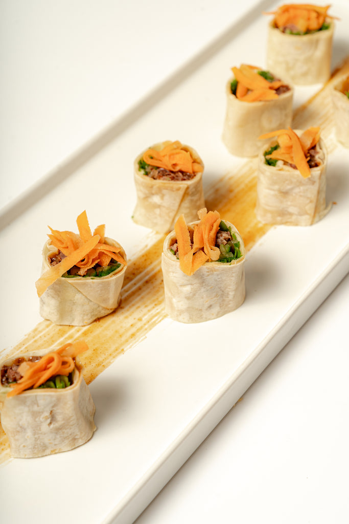 Eight Peking Duck Roulade bites, filled with greens, shredded carrots, and scallions, are arranged on a white rectangular plate with a streak of hoisin sauce underneath.