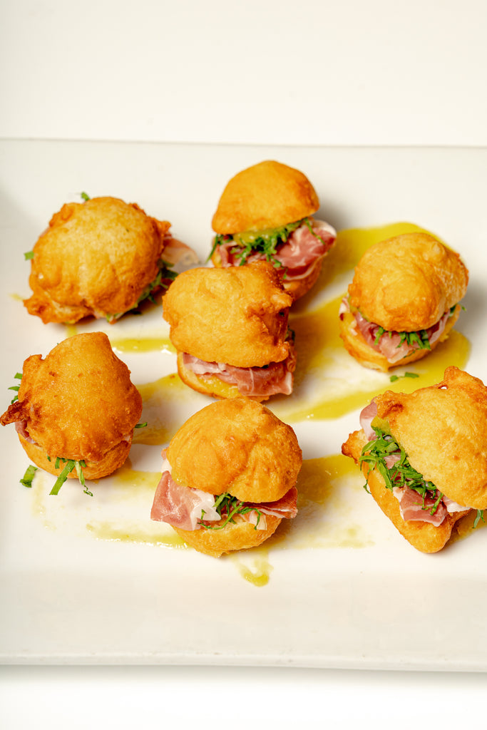 Six Savory Zeppole Bites—golden, fried buns filled with prosciutto and baby arugula—are arranged on a white plate and drizzled with olive oil.