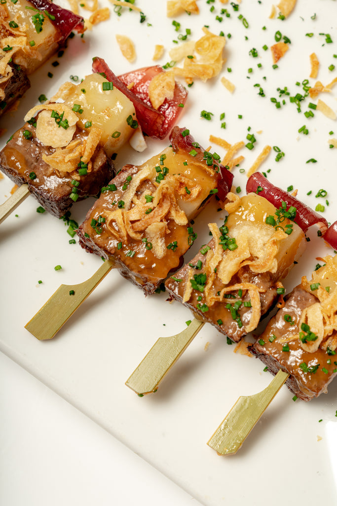 Four Short Rib Kebab skewers of grilled meat and vegetables, topped with crispy fried onions, chopped chives, and tangy pickled red onion, arranged on a white plate.
