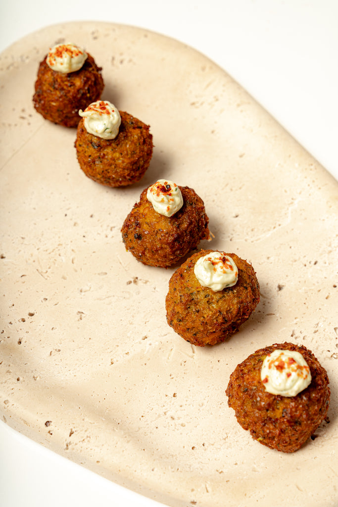 Five Yam Falafel balls topped with citrus raita and sprinkled spices, arranged diagonally on a beige ceramic platter.